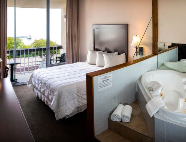 A spacious Yorktown hotel room with a white linen bed, large jacuzzi, and a glass sliding door to a private balcony with water front view.