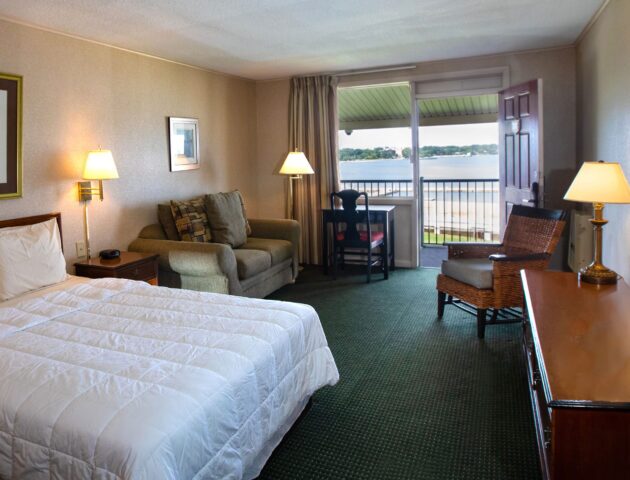 A spacious Yorktown hotel room with colonial green carpet, a queen bed, a love seat, desk, and terrace.