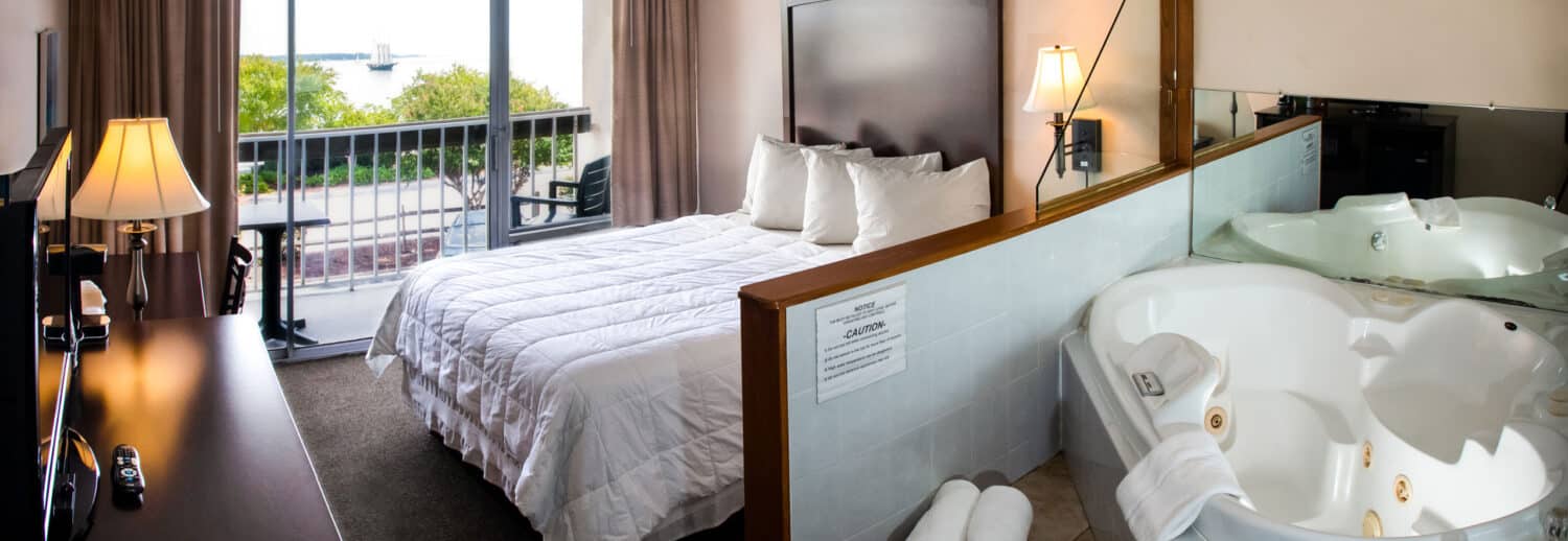A spacious Yorktown hotel room with a white linen bed, large jacuzzi, and a glass sliding door to a private balcony with water front view.