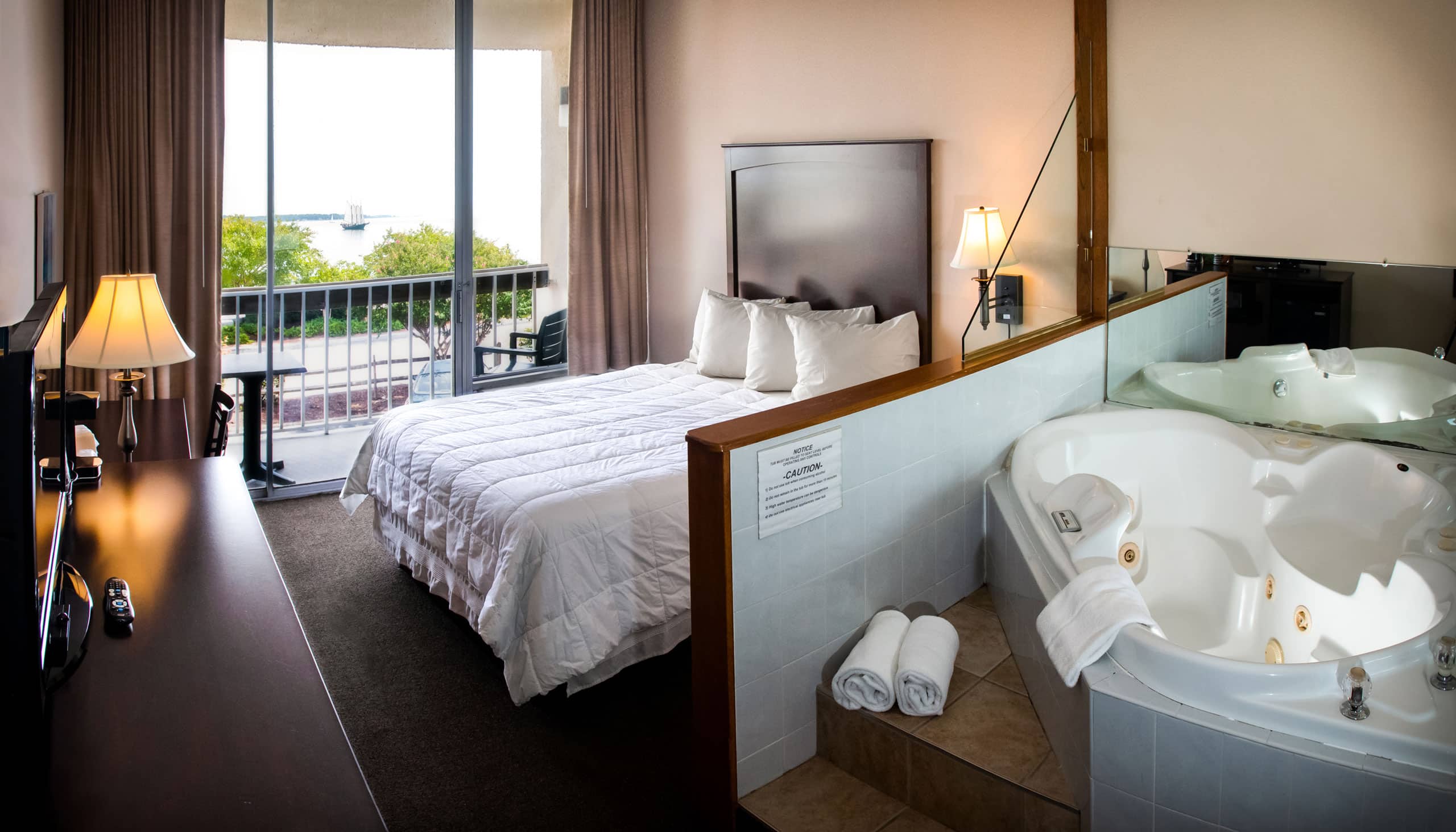 A spacious Yorktown hotel room with a white linen bed, large jacuzzi, and a glass sliding door to a private balcony with water front view.