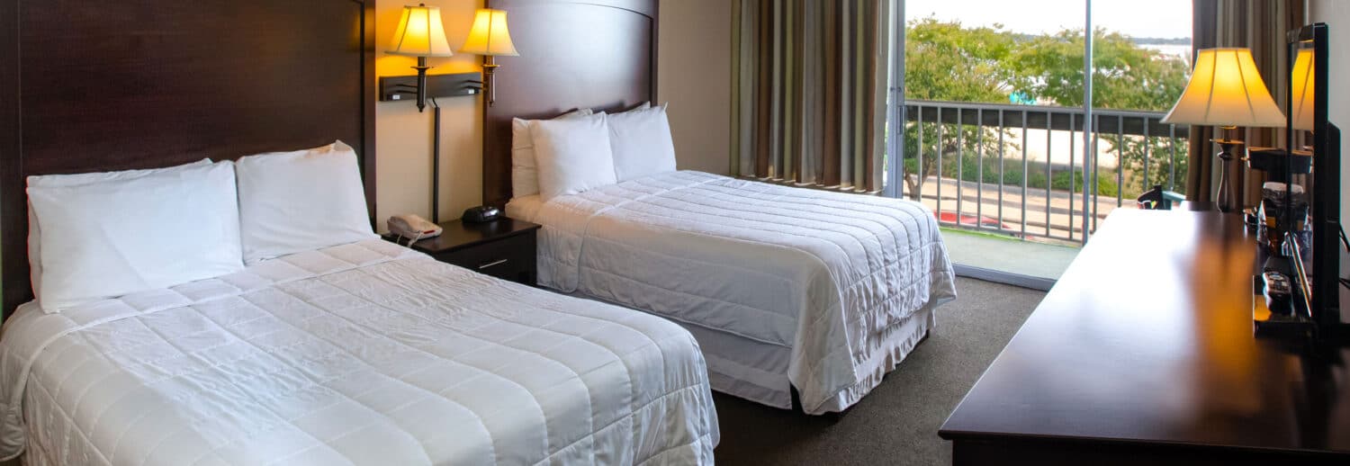 A spacious Yorktown hotel room with two full-sized beds, white linens, and a glass sliding door to a private balcony in Yorktown.