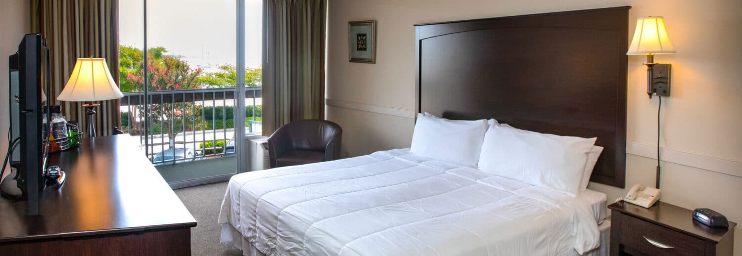 A spacious Yorktown hotel room with a large wooden headboard, white linens, and glass sliding door to a private balcony in Yorktown.