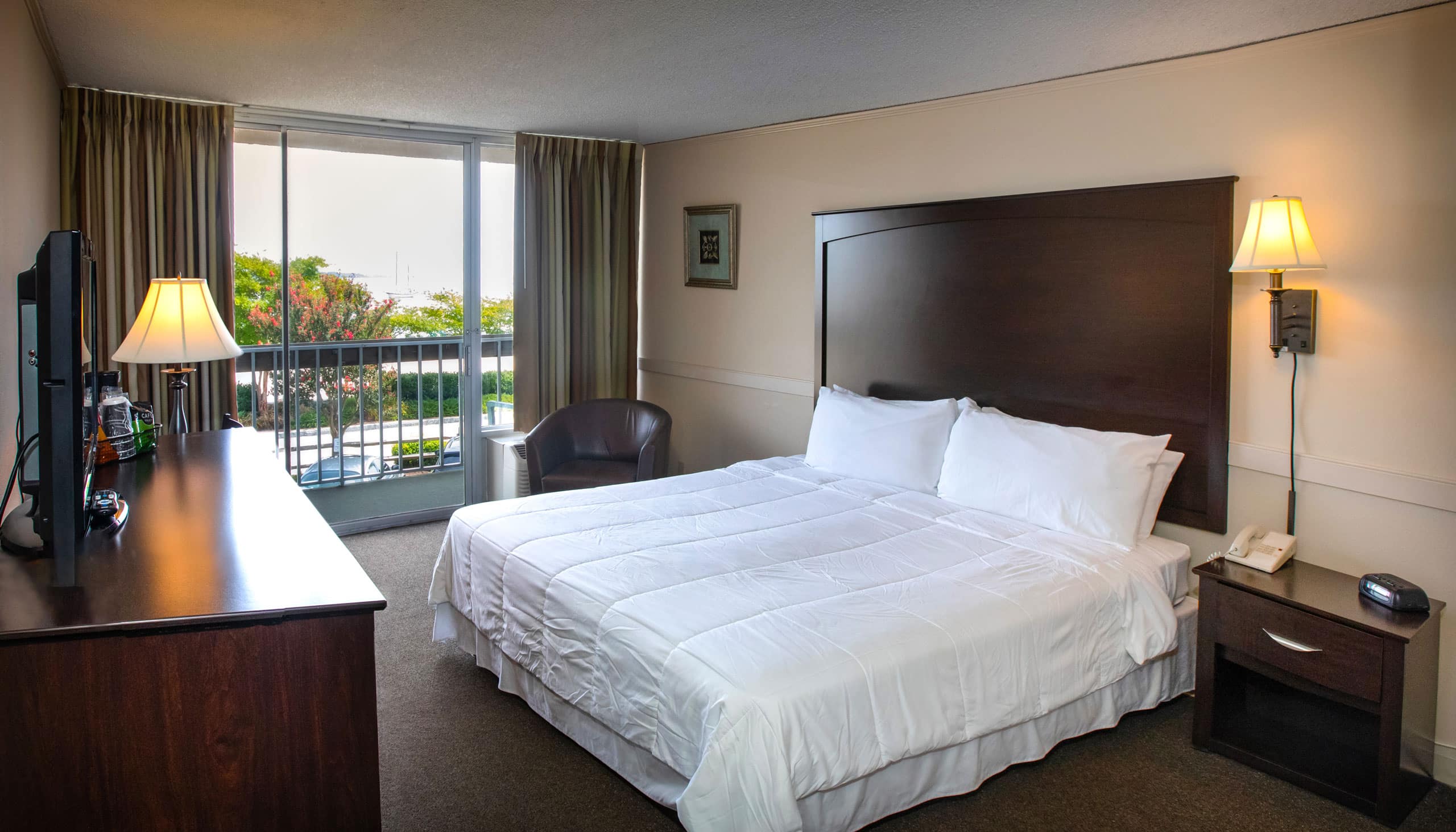 A spacious Yorktown hotel room with a large wooden headboard, white linens, and glass sliding door to a private balcony in Yorktown.
