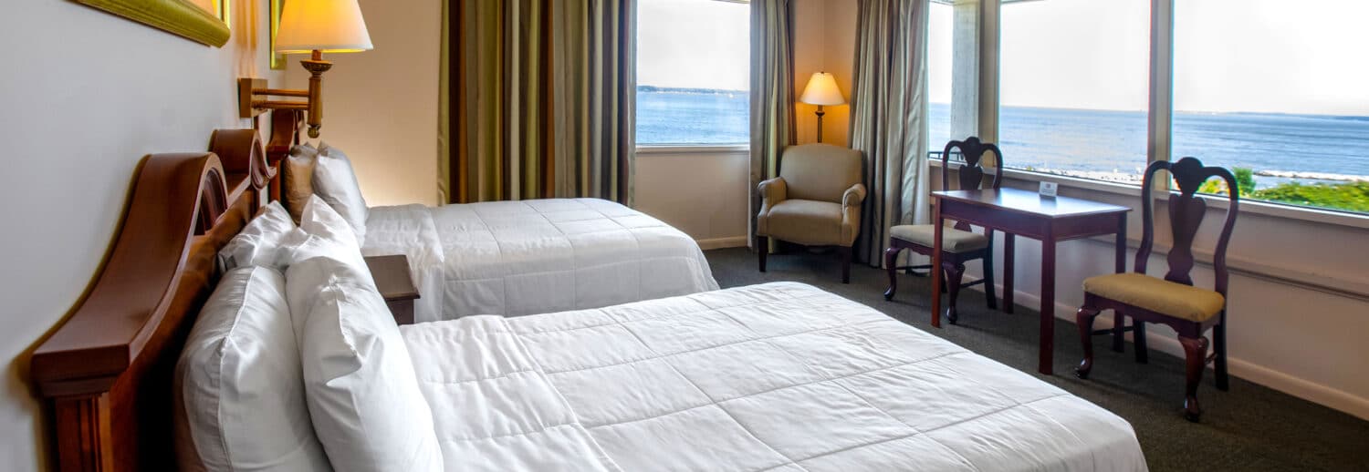 A spacious Yorktown hotel room with colonial green carpet, two queen beds with white linens, a desk, and expansive waterfront views.