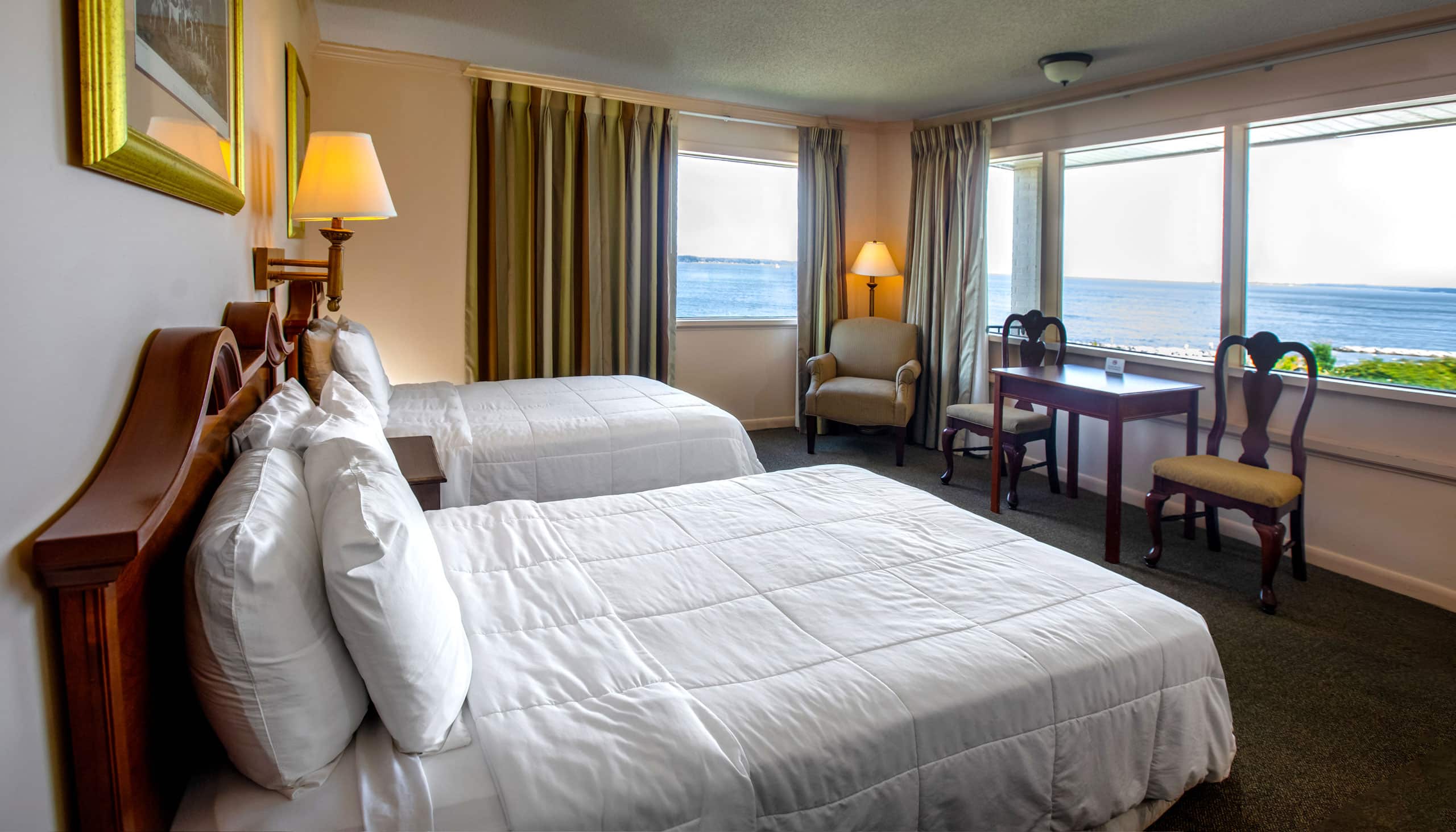 A spacious Yorktown hotel room with colonial green carpet, two queen beds with white linens, a desk, and expansive waterfront views.