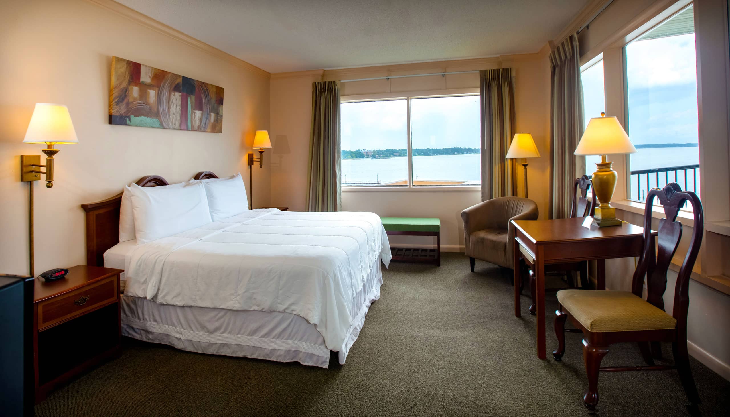 A spacious Yorktown hotel room with colonial green carpet, a king bed with white linens, a desk, and waterfront views.