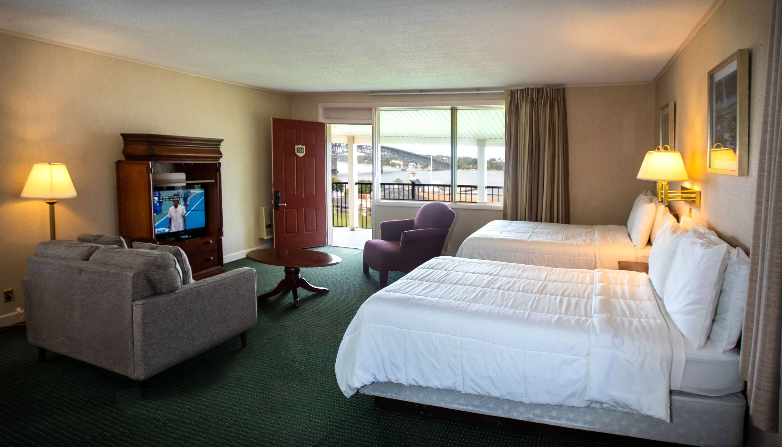 A spacious Yorktown hotel room with colonial green carpet, two full beds, a sofa set, and a TV.