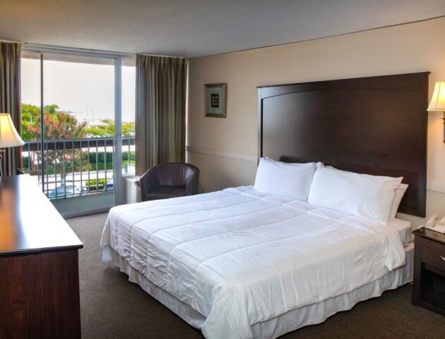A spacious Yorktown hotel room with a large wooden headboard, white linens, and glass sliding door to a private balcony in Yorktown.